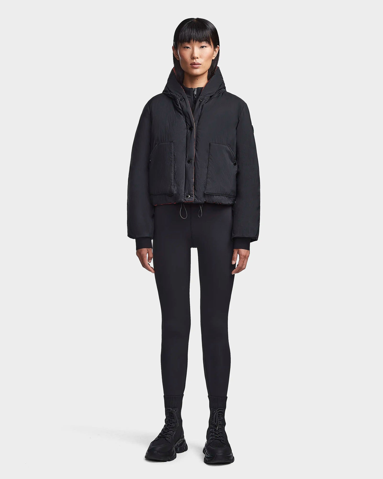 A person with straight black hair faces forward, wearing the Rudsak BALA - 8123614 Orange sustainable puffer women’s winter coat, black leggings, and black shoes against a plain white background.