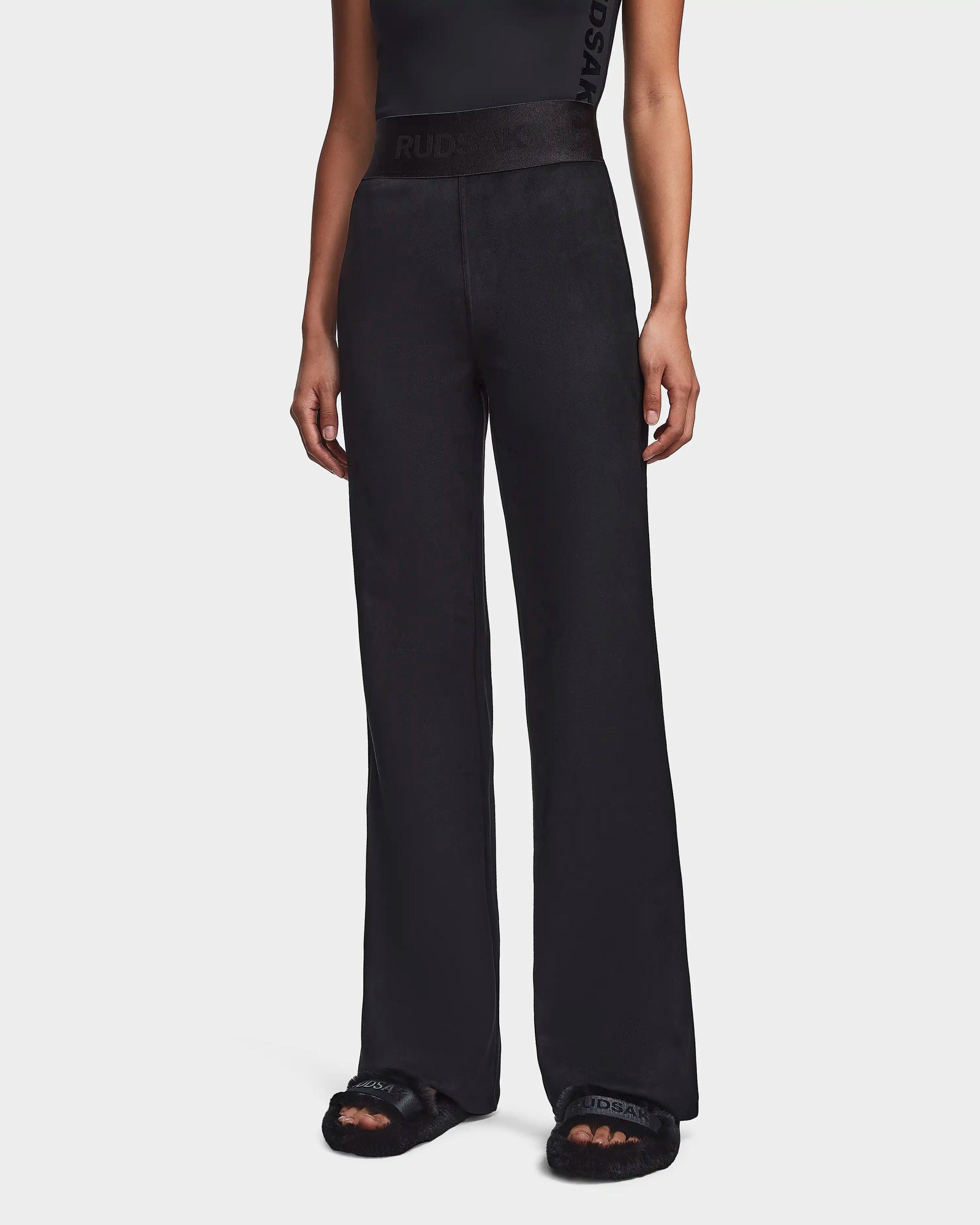 A person wears the Rudsak SEDONA - 8523541 Black high-waisted, flare leg pants and black fuzzy slippers with a sleeveless top. The photo captures this casual chic look from shoulders to feet.