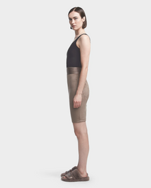 A woman stands in profile against a plain background, wearing the Rudsak CHEYNA - 8523514 Cement skirt with a fitted silhouette and elasticized waistband, paired with a black sleeveless top and brown fuzzy slide sandals.