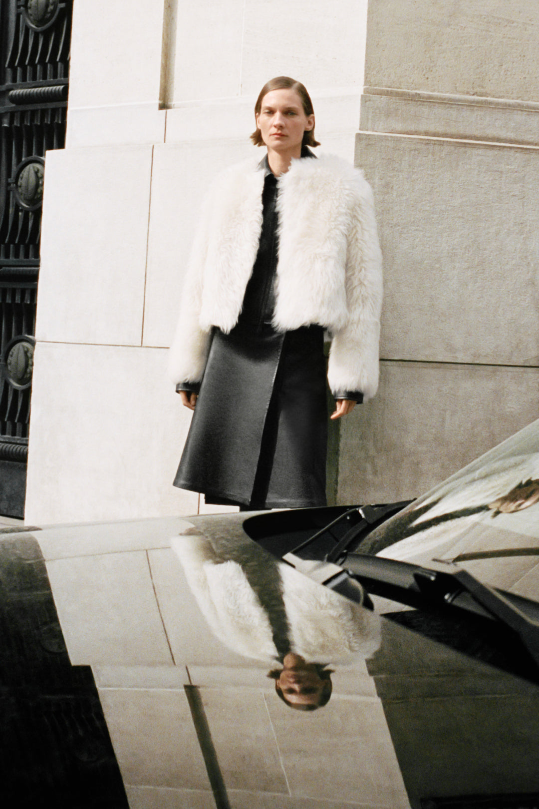 Woman wearing cream-colored shearling jacket over black outfit against urban stone backdrop.