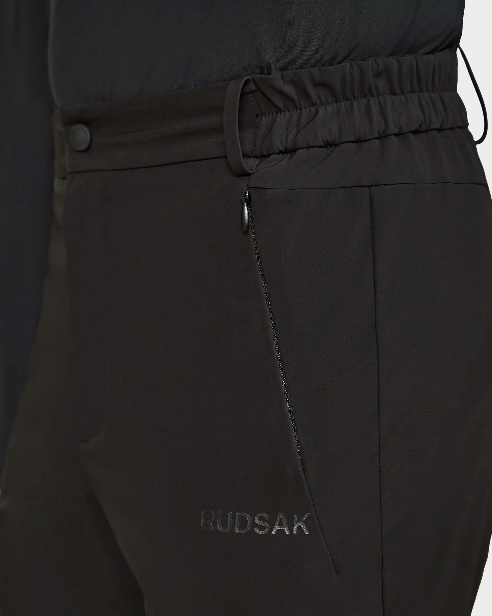 Close-up of Rudsak KICO - 6523508 Black tailored men’s outdoor pants with an elastic waistband, zippered side pocket, and subtle branding by the pocket, paired with a matching dark top.