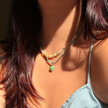 Shimmering beaded necklace made of smooth opal rondels in shades of tan, teal, orange and pink on a gold linking ovals clasp. Styled on a neck layered with a turquoise slim gemstone and a acorn drop charm.