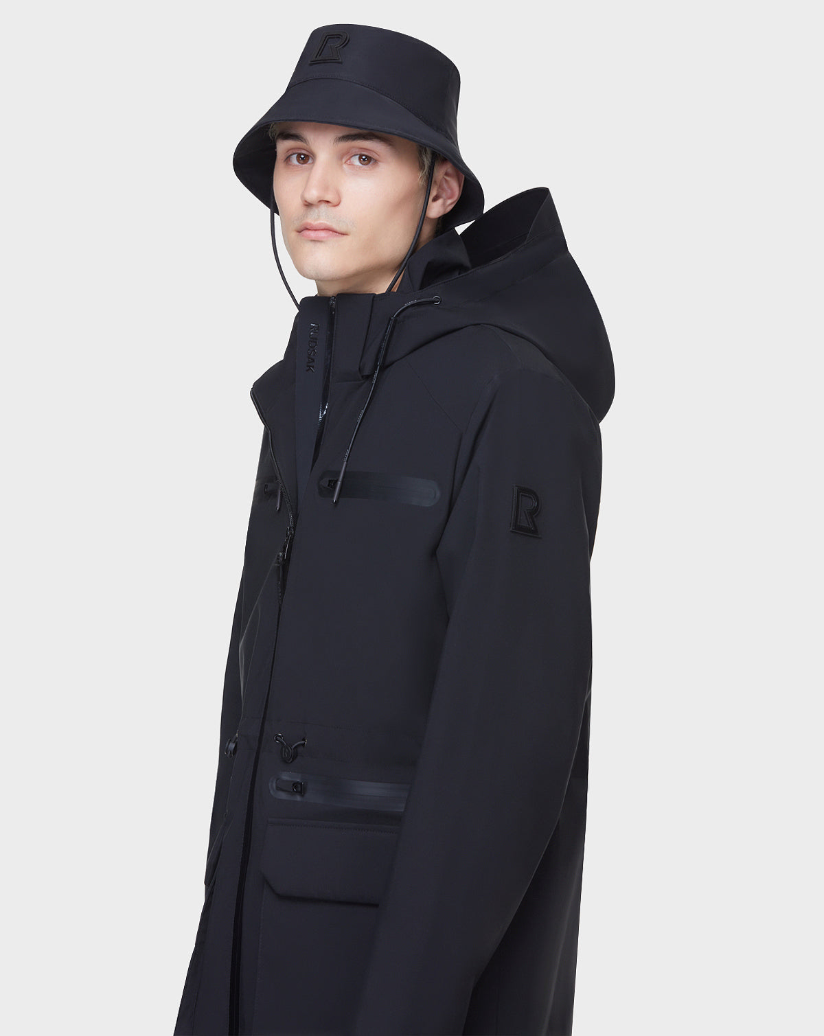 A person in RUDSAK’s JACY - 6122000 BLACK hooded jacket and black bucket hat faces the camera. The minimalistic look, with subtle logo patches, is inspired by classic men’s rain trench coats. Plain white background.