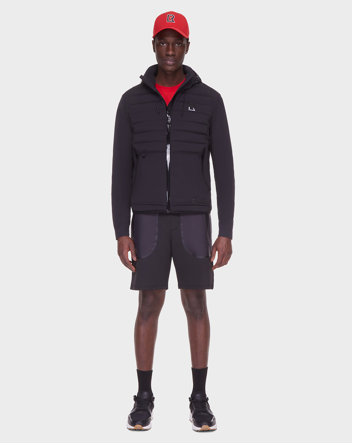 A man faces forward on a plain white background wearing the RUDSAK JAS - 6122006 BLACK men’s windbreaker with RDS-certified down, paired with a red cap, red shirt, black shorts with gray pockets, black socks, and black sneakers.