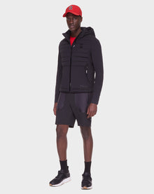 A man with a neutral expression stands against a white background wearing the RUDSAK JAS - 6122006 BLACK mens windbreaker, paired with black shorts, black sneakers, and a red baseball cap; his hands rest at his sides.