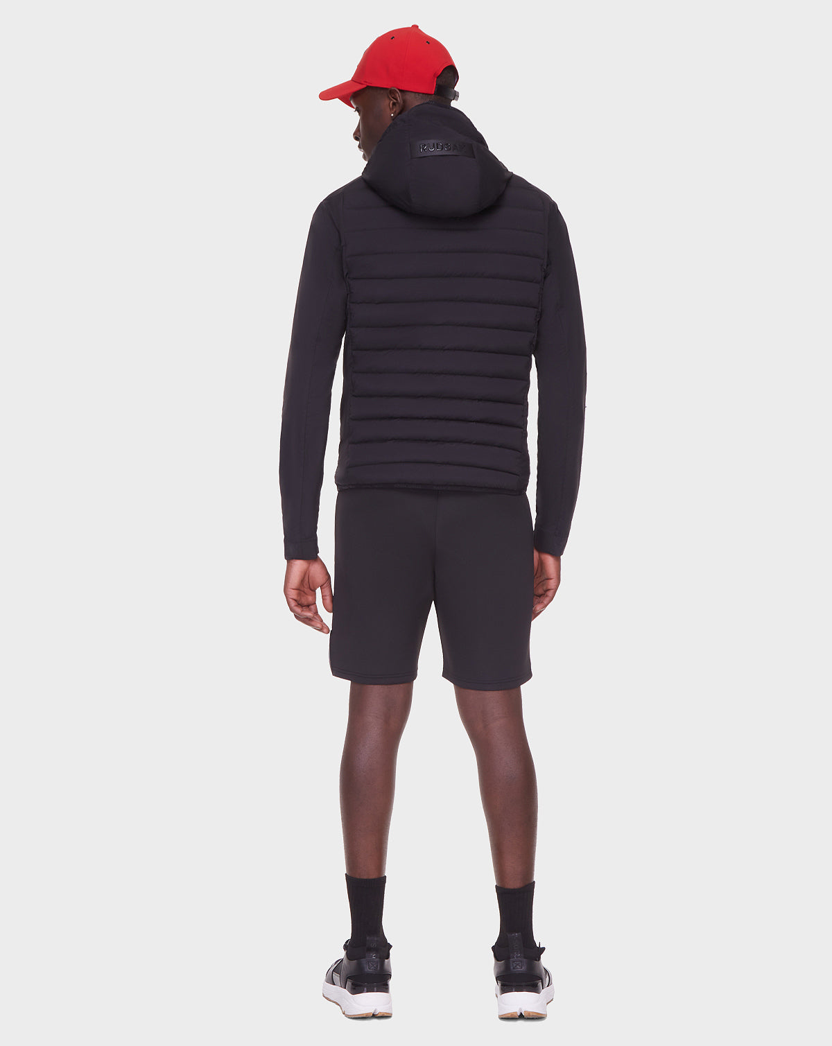 A person facing away wears the RUDSAK JAS - 6122006 BLACK men’s windbreaker, paired with black shorts, black socks, black and white sneakers, and a red cap against a plain white background.