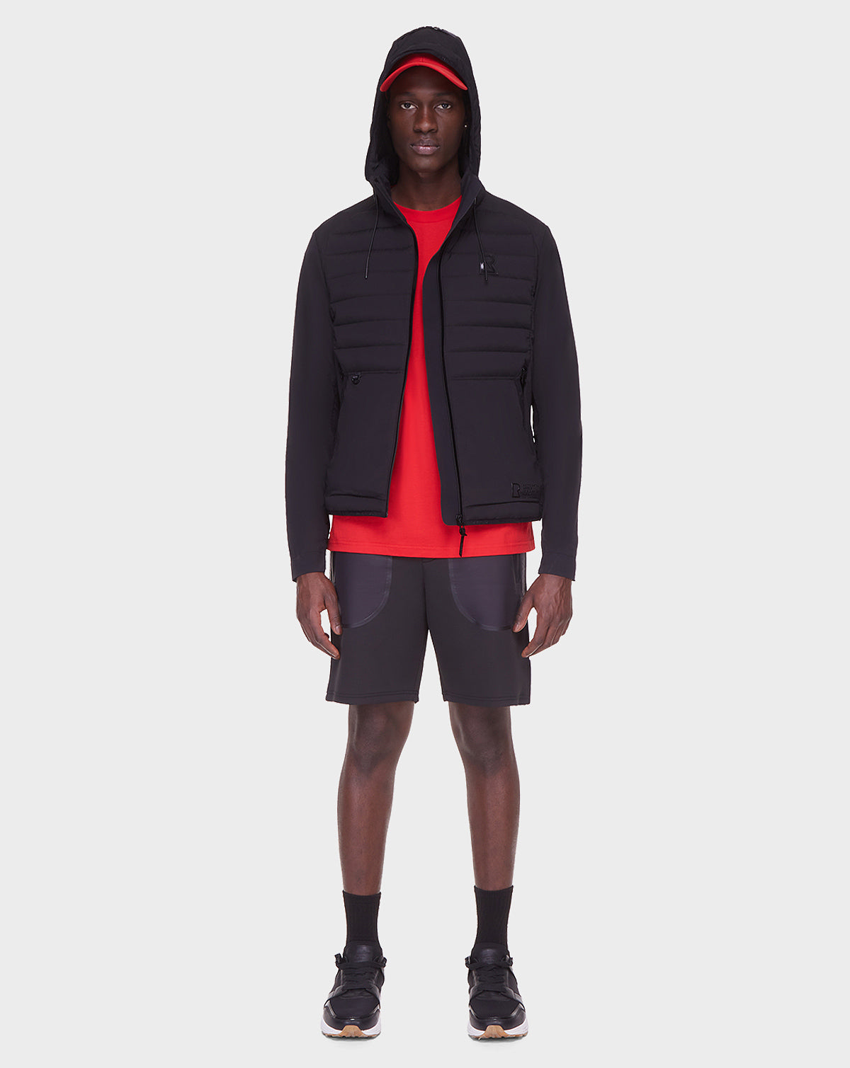 A man faces forward against a plain white background, wearing the RUDSAK JAS - 6122006 BLACK men’s windbreaker with lightweight stretch fabric, a red shirt and cap, black shorts, socks, and sneakers.