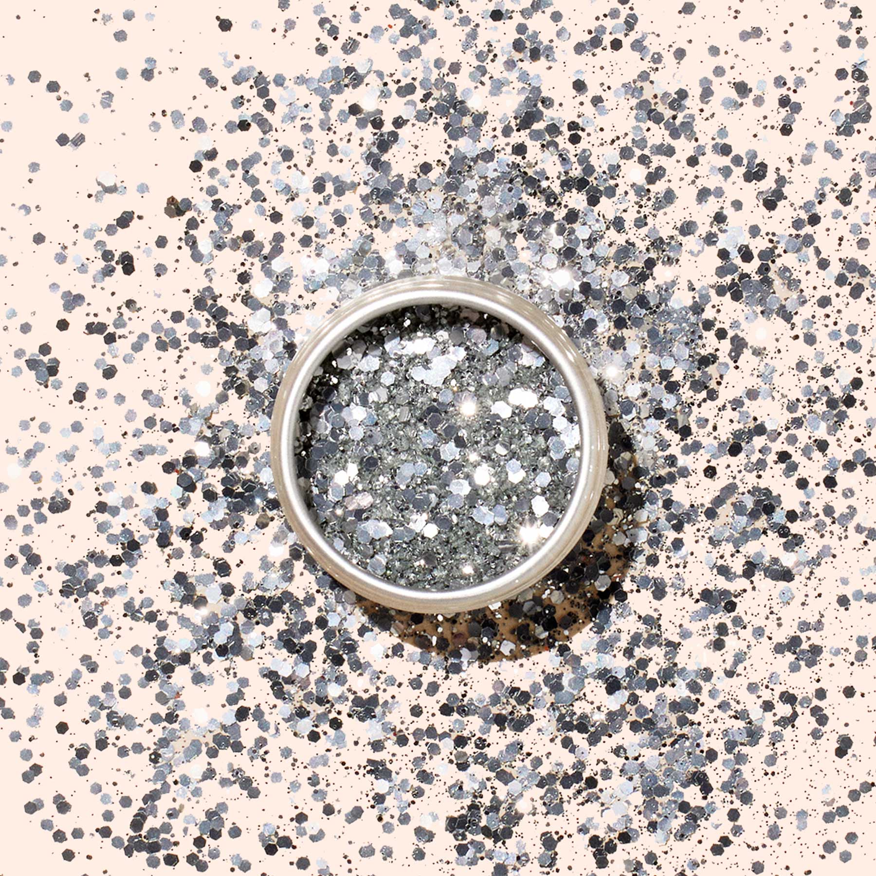 A round container of Manucurist Glitters Duo Silver  Mirror is displayed on a light surface, with its silver and mirror glitters shimmering in a circular pattern around it.