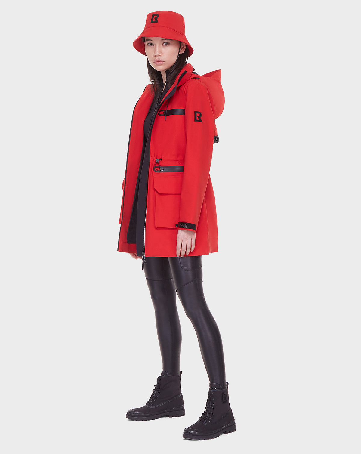 A woman stands sideways against a white background wearing the Rudsak CAMY - 8122002 POPPY RED rain trench with removable hood, paired with a matching red bucket hat, black leggings, and black boots.