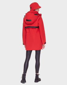 A person faces away, wearing the Rudsak CAMY - 8122002 POPPY RED water-resistant coat with removable hood, a matching red bucket hat, black shiny leggings, and black lace-up boots against a plain white background.