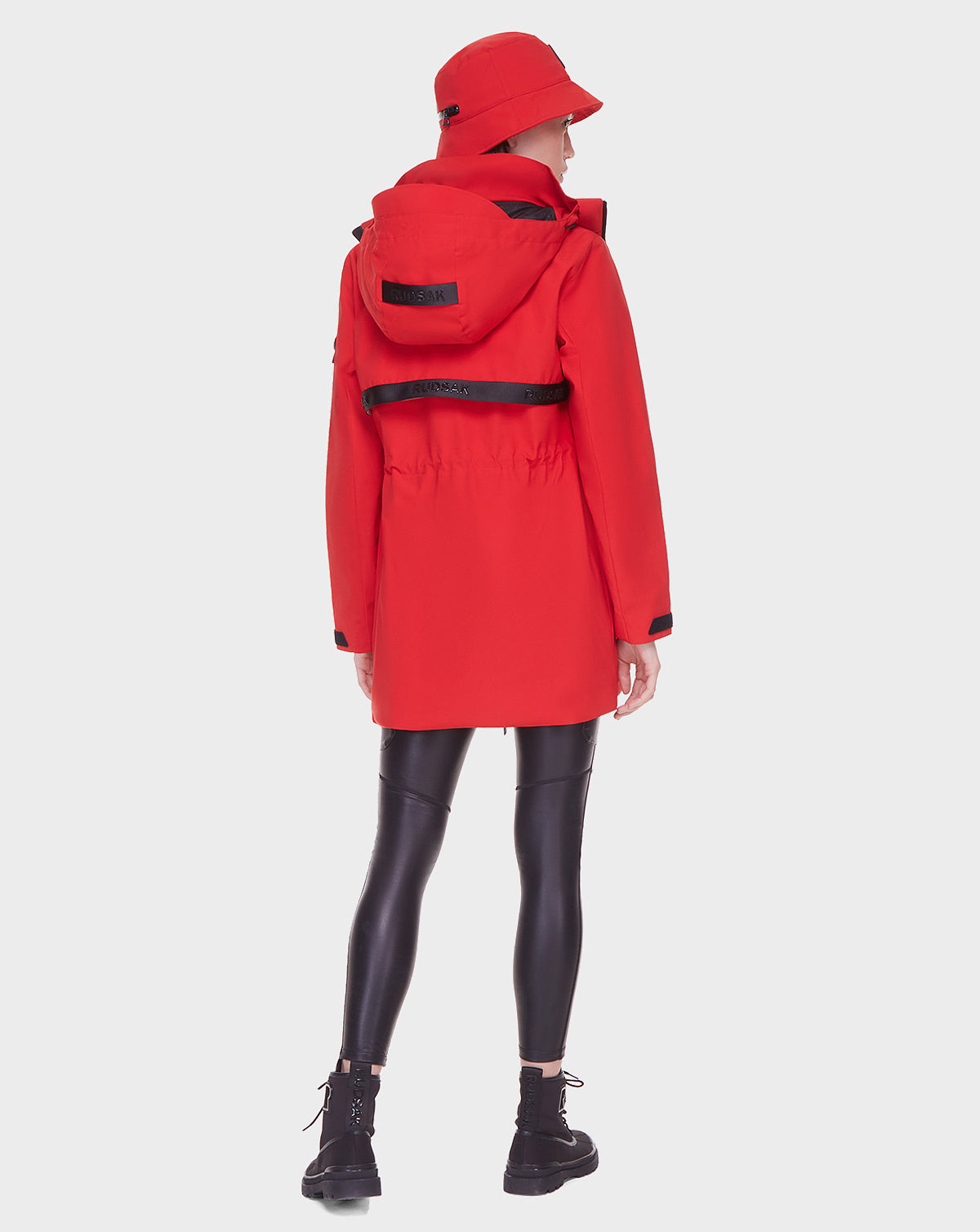 A person faces away, wearing the Rudsak CAMY - 8122002 POPPY RED water-resistant coat with removable hood, a matching red bucket hat, black shiny leggings, and black lace-up boots against a plain white background.